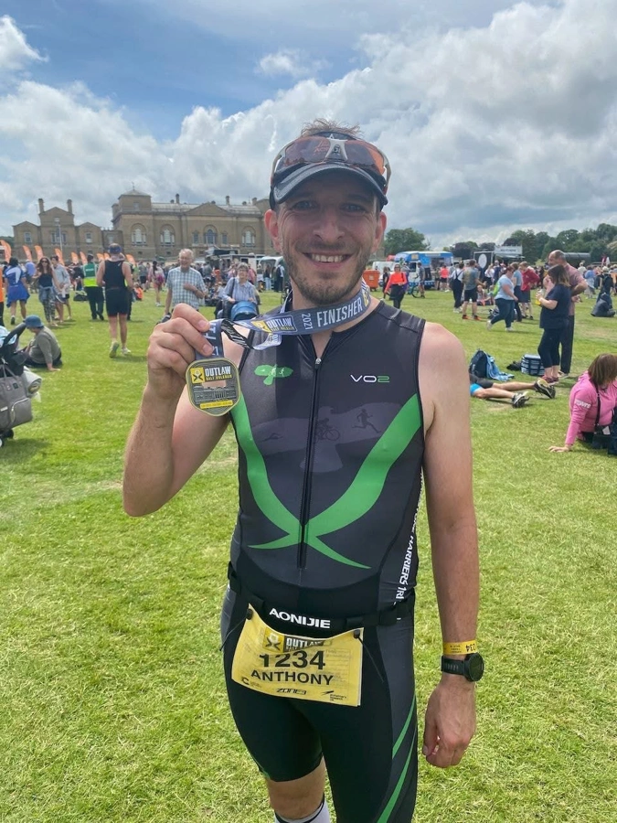 Anthony Warren holding his Outlaw Half finisher medal in the event village, smiling in Maidstone Harriers tri suit