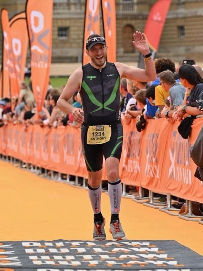 Anthony Warren celebrating on the orange finish carpet at an Outlaw Triathlon event, wearing Maidstone Harriers tri kit.