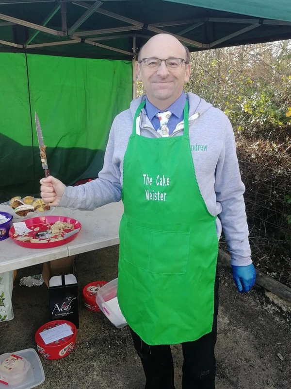 Maidstone Harriers runner Andrew Burgess wearing a green The Cake Meister apron and holding a cake knife at a running event cake table
