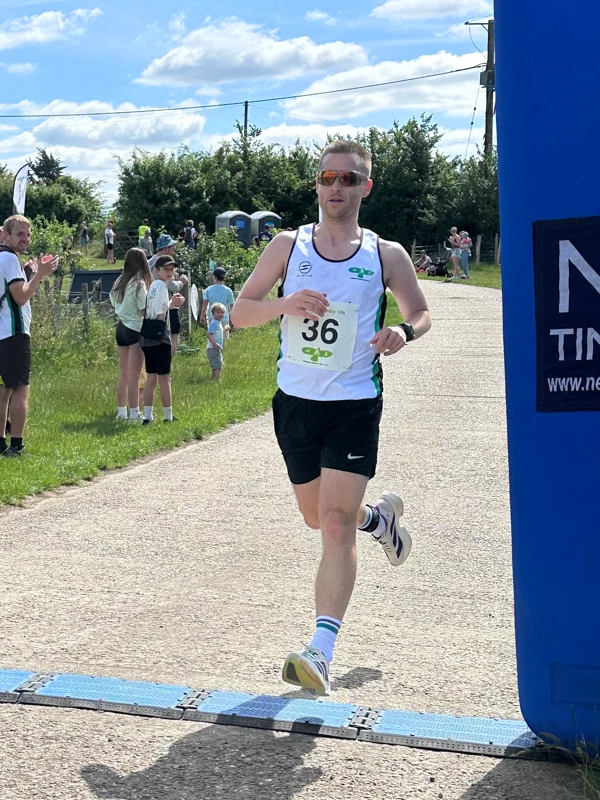 Matt Cox sprinting past the blue timing arch to claim 1st place overall at the Maidstone Harriers David Thornby 10k run, wearing the club's white running vest, black shorts, and sunglasses.