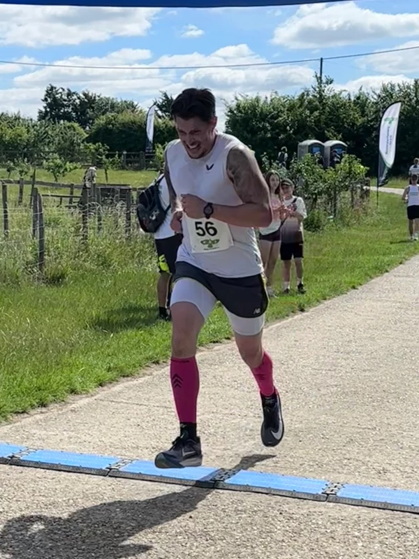 Jamie Forman pushing hard to finish in 2nd place for men at the David Thornby 10k running event, sporting a white top, grey shorts with pink compression socks, and race bib number 56.