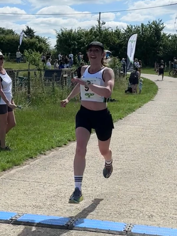 Sian Knight-James smiling as she approaches the blue timing mats to secure 2nd place for women at the Maidstone Harriers David Thornby 10k event, dressed in the club's white vest and black running shorts.