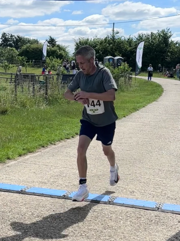 Ben Sharman checking his running watch as he crosses the finish line mat to take 3rd place at the Maidstone Harriers David Thornby 10k event, wearing a grey technical t-shirt and dark shorts.