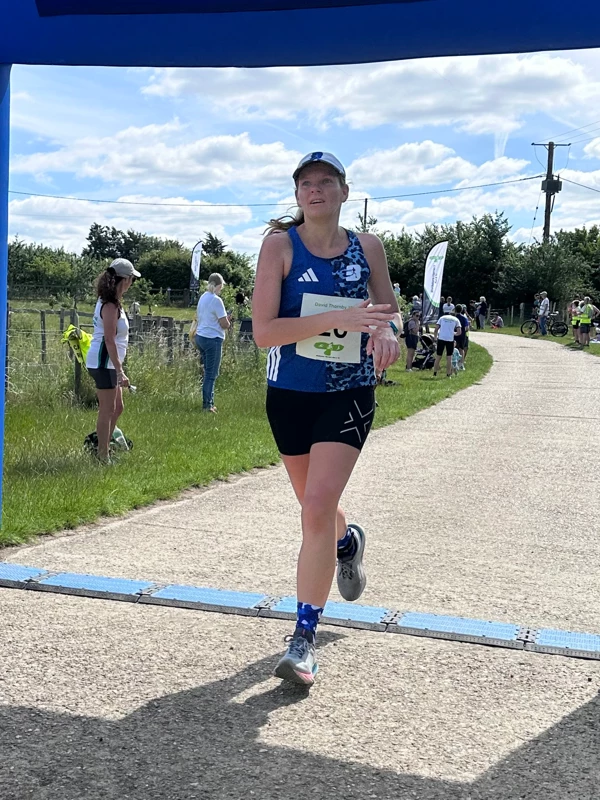 Petra Cairns crossing the finish line on a sunny day to win 3rd place in the women's race at the Maidstone Harriers David Thornby 10k at Little Fant Farm, wearing a blue running vest and black shorts.