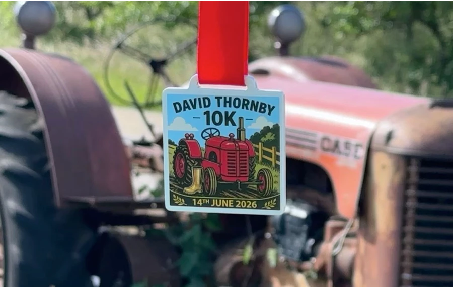 The farm-themed square finisher medal for the Maidstone Harriers David Thornby 10k race held on 14th June 2026, featuring an illustration of a red tractor and a yellow Wellington boot at Little Fant Farm, with a blurred vintage Case tractor in the background.